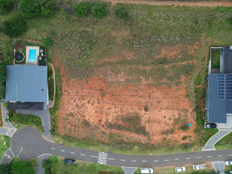 An aerial view of a vacant residential plot situated between existing developed properties with direct access to a paved estate road.