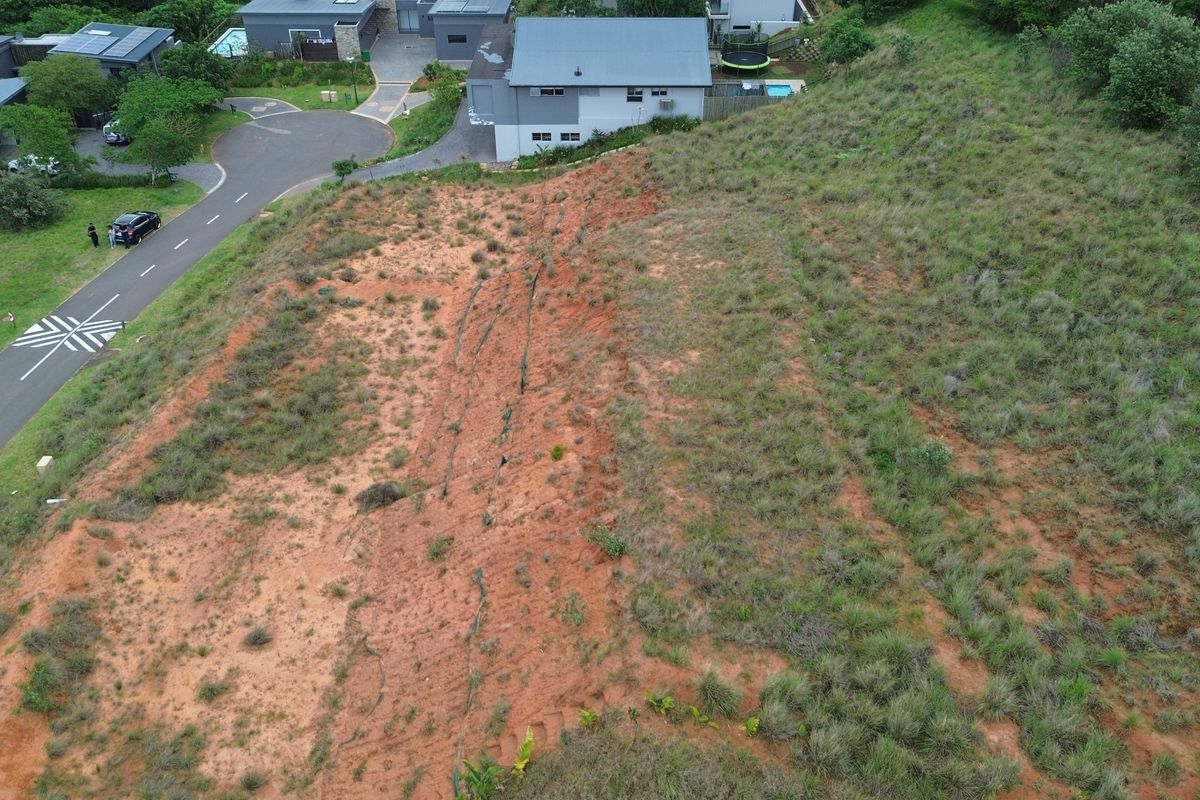 Aerial view of a vacant residential plot featuring sloped terrain and natural vegetation, situated adjacent to an established residential street.