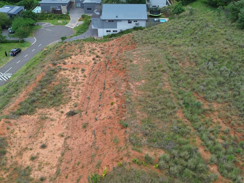 Aerial view of a vacant residential plot featuring sloped terrain and natural vegetation, situated adjacent to an established residential street.