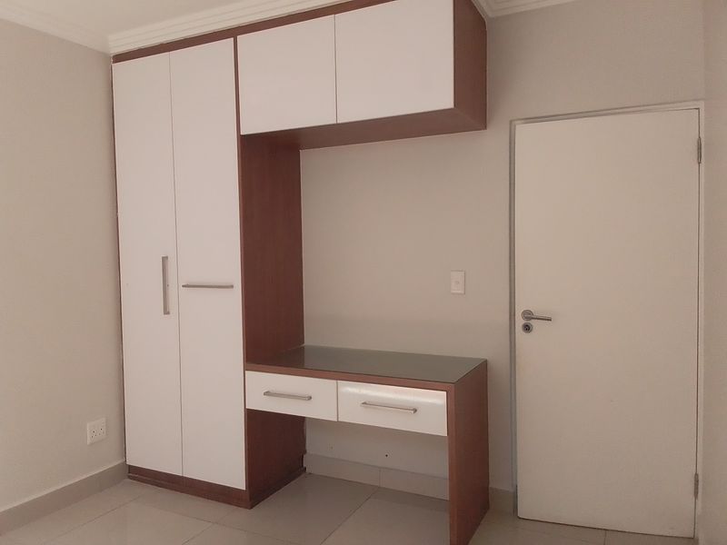A clean bedroom featuring a modern built-in wardrobe with an integrated desk and overhead storage, finished with neutral tones and tiled flooring.