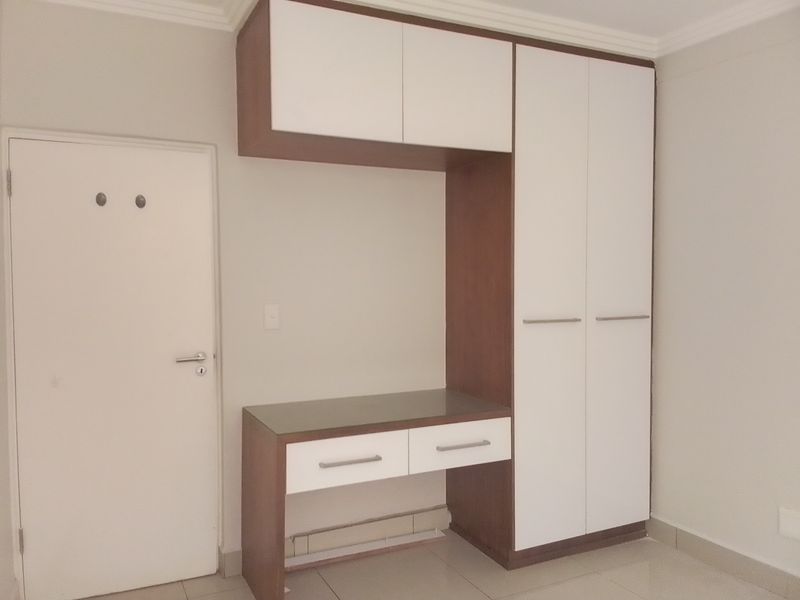 A clean bedroom featuring a modern built-in wardrobe with integrated desk space, overhead storage, and neutral tiled flooring.