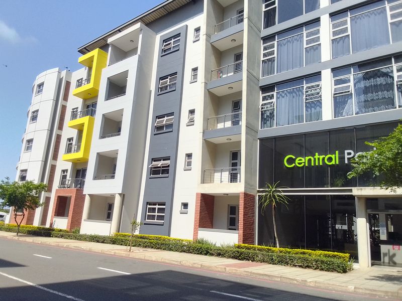 Modern multi-story apartment building facade featuring balconies with metal railings, large glass windows, and a well-maintained landscaped entrance area.