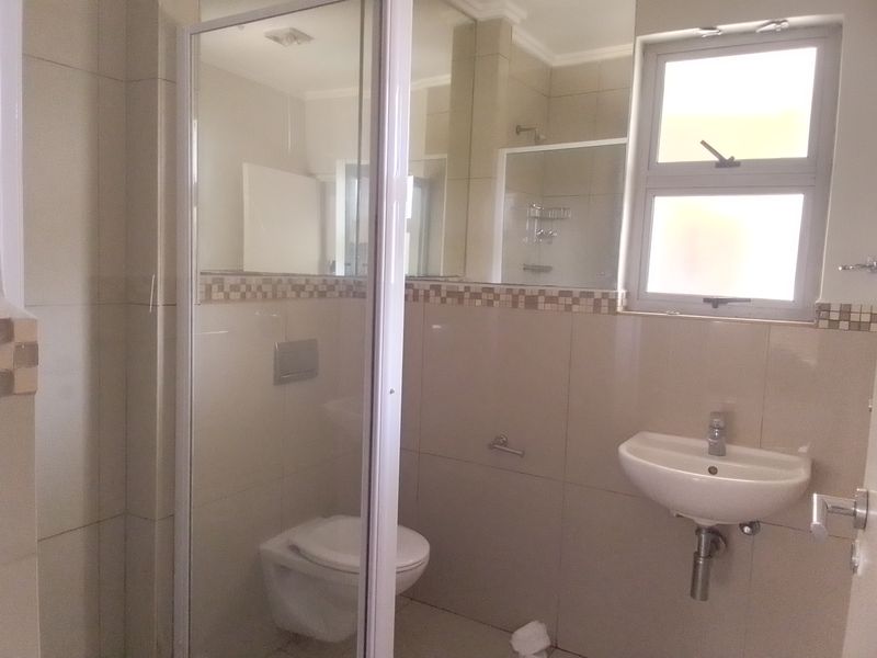 A clean, modern bathroom featuring a wall-mounted toilet, wall-hung basin, and a glass-enclosed shower with neutral wall tiling and a decorative mosaic border.