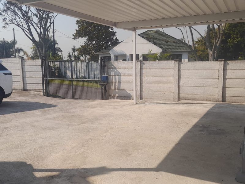 A paved driveway area featuring a covered carport and secure perimeter walling with a metal entrance gate.