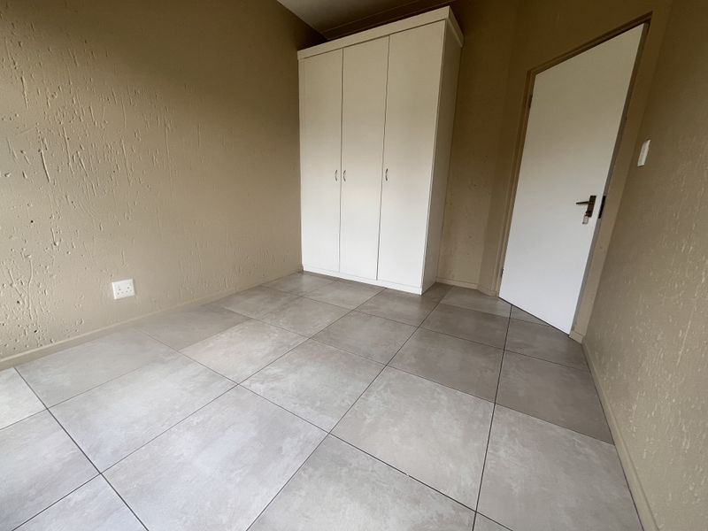 A clean, empty bedroom featuring neutral textured walls, large format grey floor tiles, and a white built-in wardrobe.