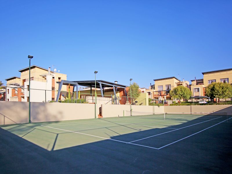 A well-maintained communal tennis court featuring perimeter fencing, floodlights, and an adjacent covered pavilion area within the apartment complex.