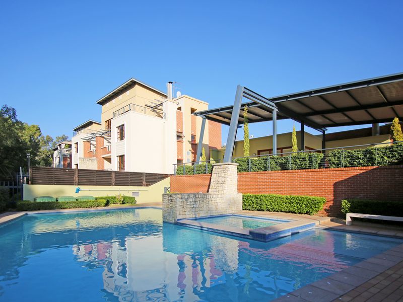 A well-maintained communal pool area featuring an integrated spa, stone water feature, and a covered patio structure set against a modern apartment complex.