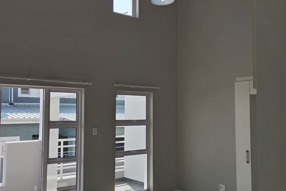 A bright, double-volume living area featuring tiled floors, exposed ceiling beams, and large windows providing access to an outdoor balcony.