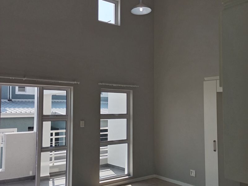 A bright, double-volume living area featuring tiled floors, exposed ceiling beams, and large windows providing access to an outdoor balcony.
