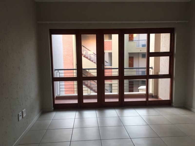 An empty living room featuring neutral tiled flooring and large timber-framed glass doors opening onto a balcony with a metal railing.
