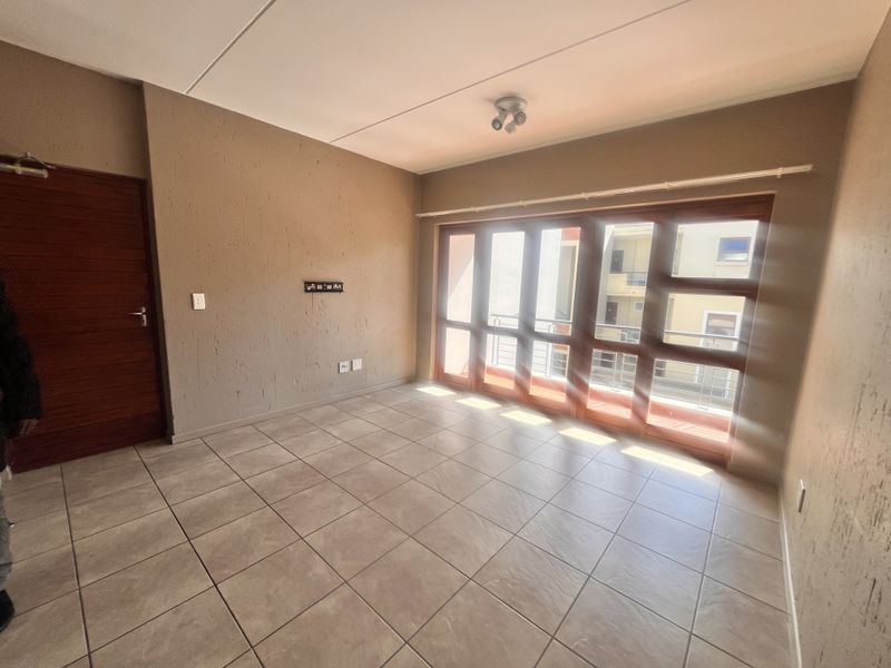 A bright, empty living room featuring neutral tiled flooring, textured walls, and large windows that provide ample natural light.