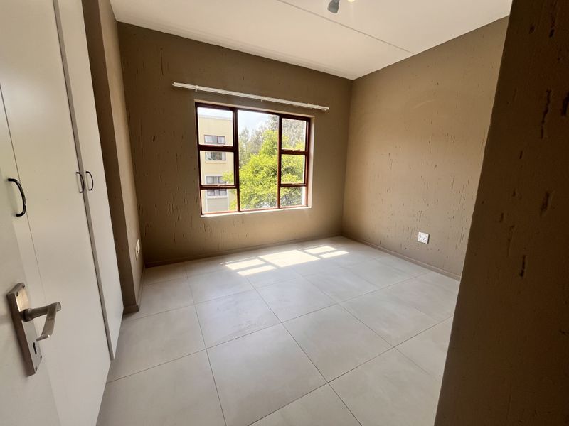 A bright, empty bedroom featuring neutral tiled flooring, built-in cupboards, and a window providing natural light and a view of the surrounding area.
