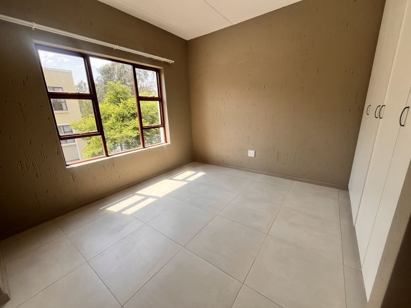 A bright, empty bedroom featuring neutral tiled flooring, built-in cupboards, and large windows allowing for ample natural light.