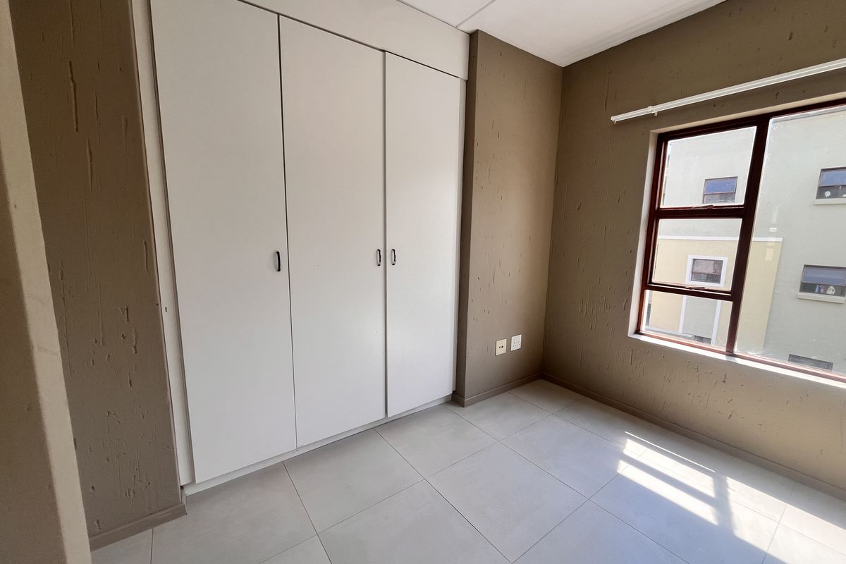 A bright, empty bedroom featuring neutral tiled flooring, textured brown walls, and white built-in cupboards with ample storage space.