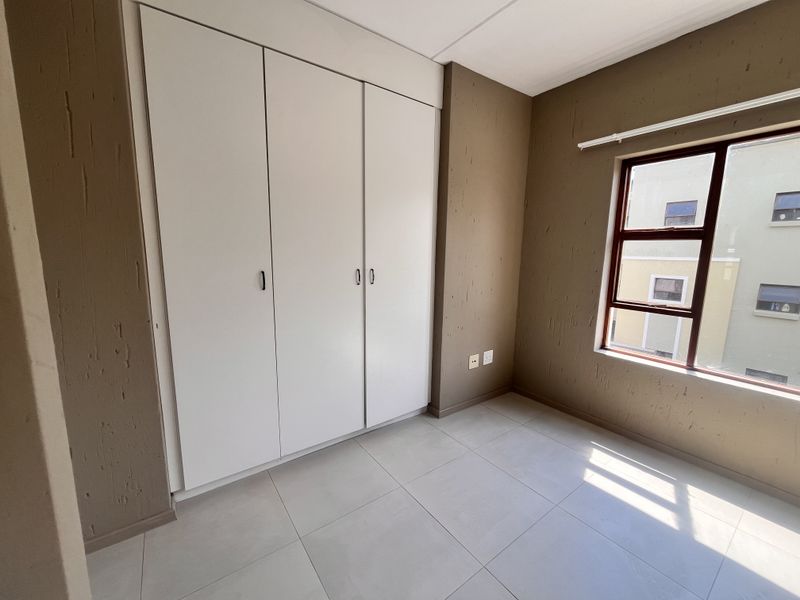 A bright, empty bedroom featuring neutral tiled flooring, textured brown walls, and white built-in cupboards with ample storage space.