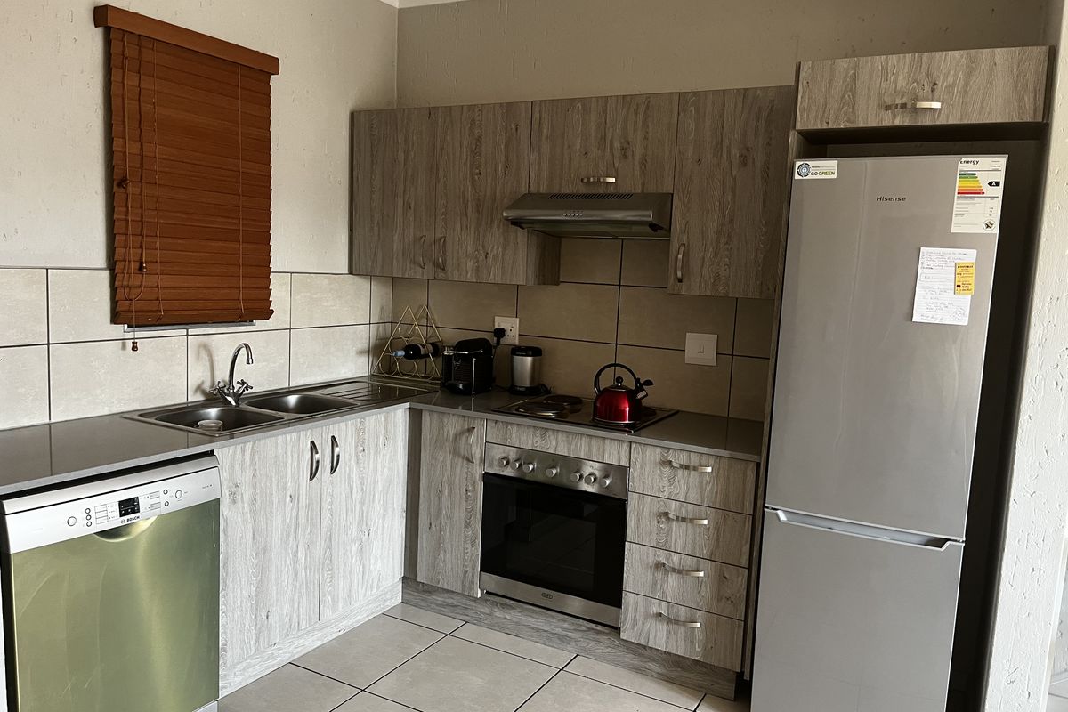 A functional kitchen featuring wood-look cabinetry, stainless steel appliances including an oven and hob, tiled backsplash, and neutral floor tiles.