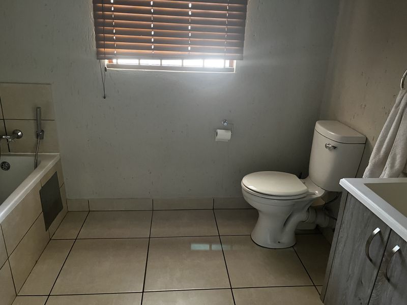 A functional bathroom featuring a built-in bathtub, toilet, and vanity cabinet with tiled flooring and window blinds.
