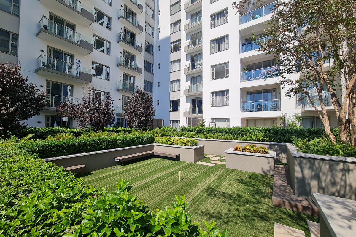 A well-maintained communal garden area featuring artificial turf, raised planters, and built-in bench seating, set against a modern apartment building with glass balconies.