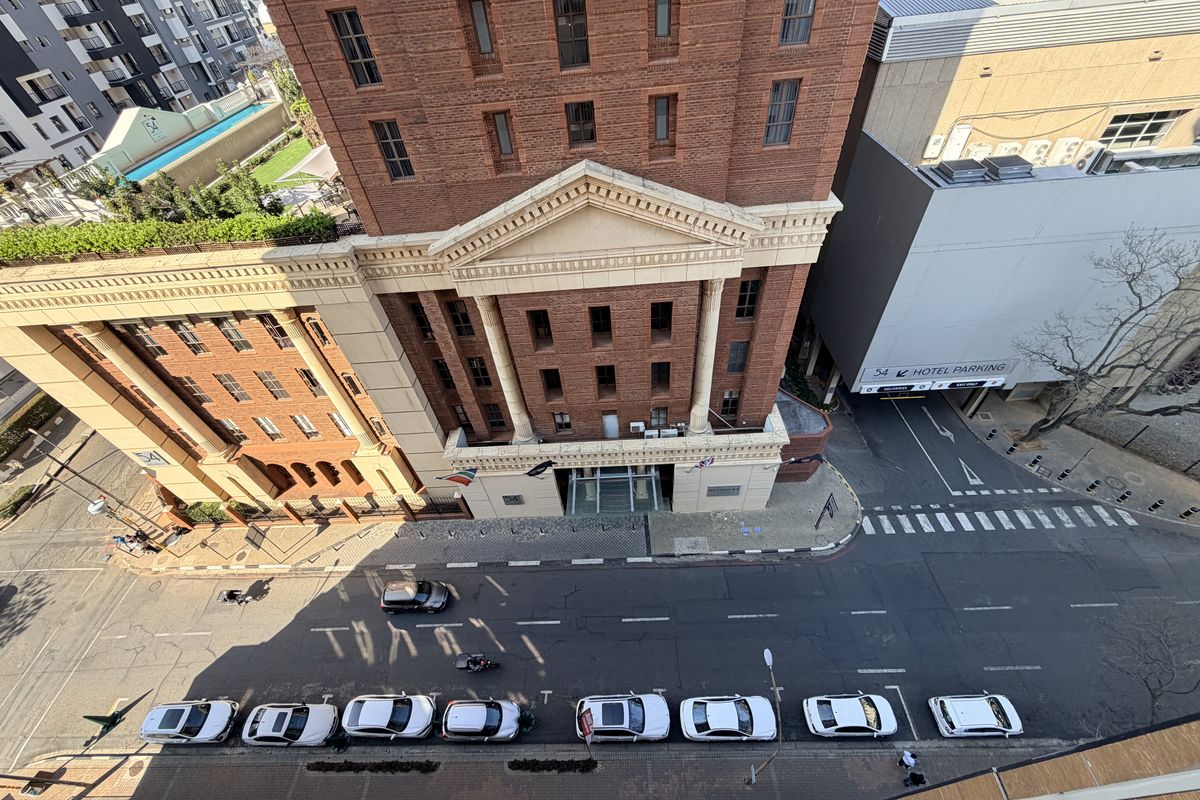 An elevated aerial view looking down onto a city street featuring a historic brick building with classical architectural details and adjacent hotel parking.