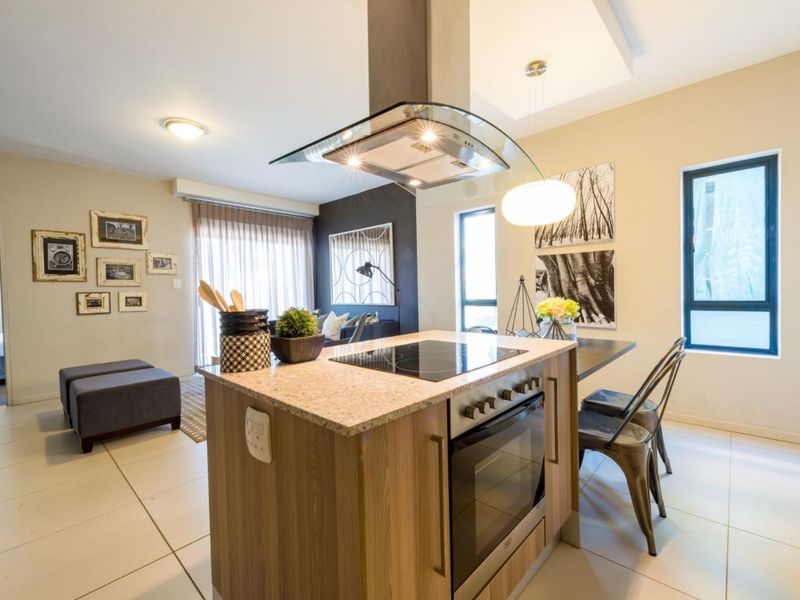 Modern open-plan living area featuring a central kitchen island with built-in oven, hob, and extractor fan, complemented by neutral tiled flooring throughout.