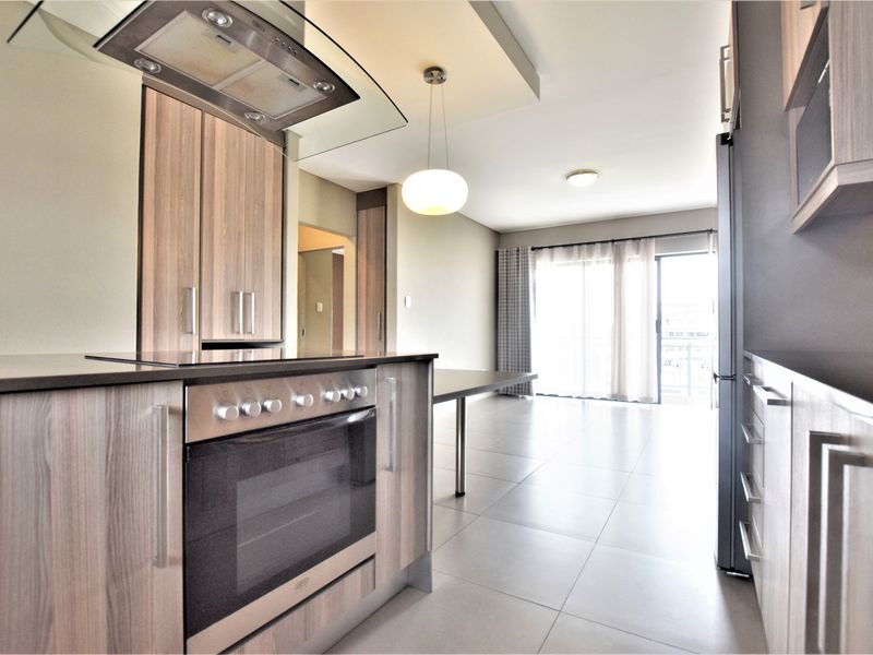 Modern open-plan kitchen and living area featuring sleek wood-finish cabinetry, built-in appliances, tiled flooring, and direct access to a balcony through sliding glass doors.