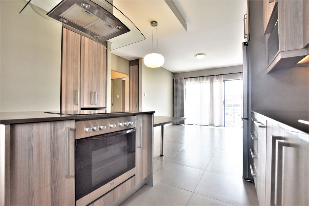 Modern open-plan kitchen and living area featuring sleek wood-finish cabinetry, built-in appliances, tiled flooring, and direct access to a balcony through sliding glass doors.