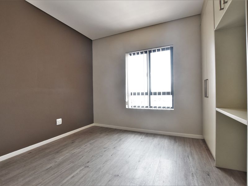 A bright, empty bedroom featuring modern laminate flooring, a neutral color palette, and a built-in wardrobe with integrated shelving.