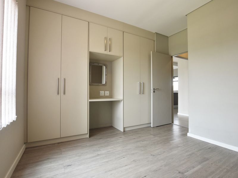 A bright, empty bedroom featuring neutral-toned built-in cupboards with an integrated vanity nook and modern laminate flooring.