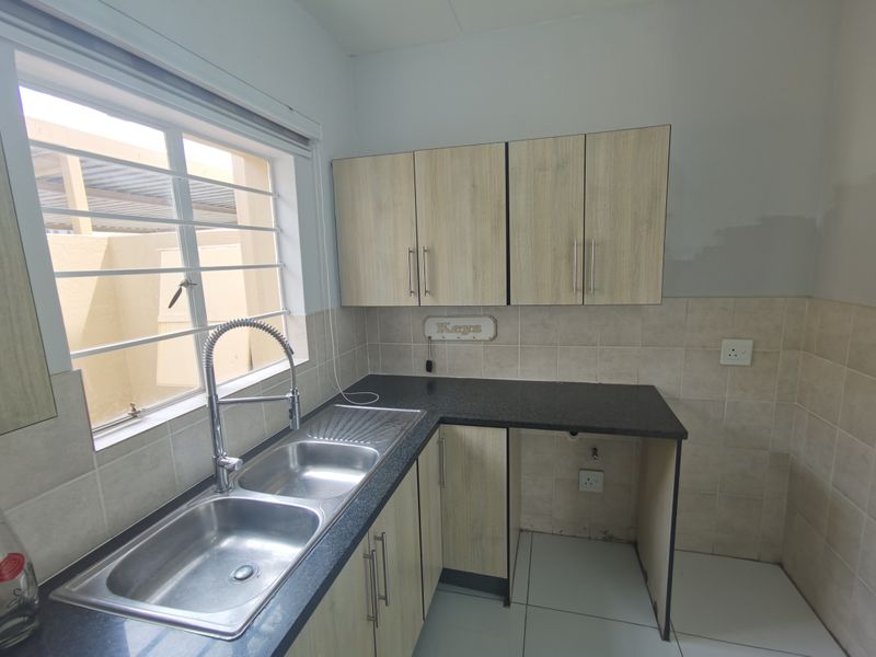 A functional kitchen area featuring light wood-finish cabinetry, granite countertops, a double stainless steel sink, and tiled splashbacks with space for an under-counter appliance.