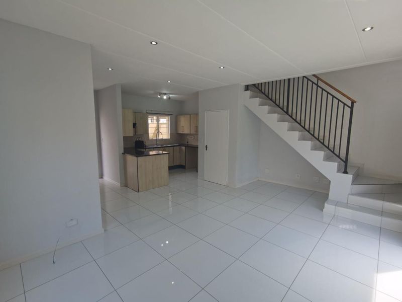 Open-plan living area featuring white tiled flooring, recessed lighting, and a staircase with a metal balustrade, leading to a compact kitchen with built-in cabinetry.
