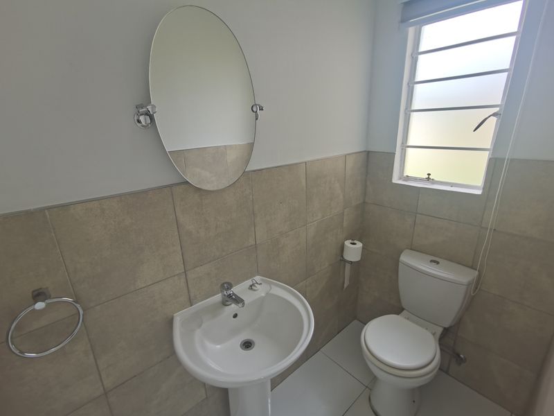 A clean and functional bathroom featuring a pedestal basin, toilet, and neutral wall tiling with a louvered window for natural light.