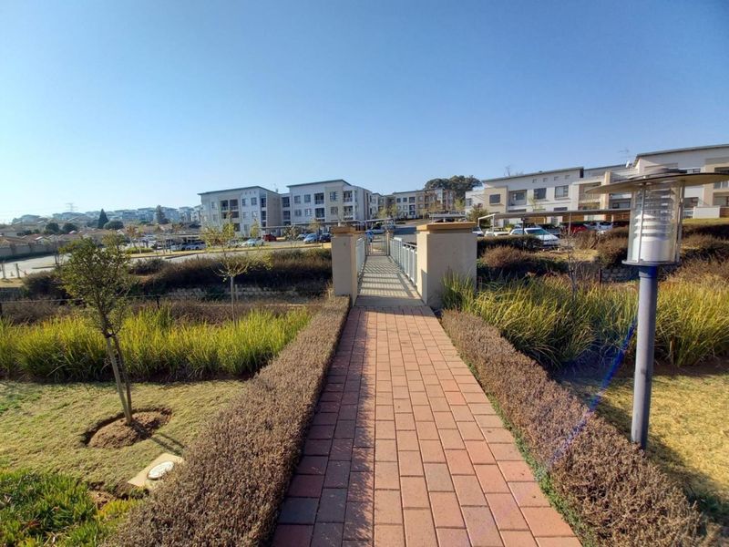 A well-maintained paved walkway leading across a pedestrian bridge through a landscaped garden area within the apartment complex grounds.
