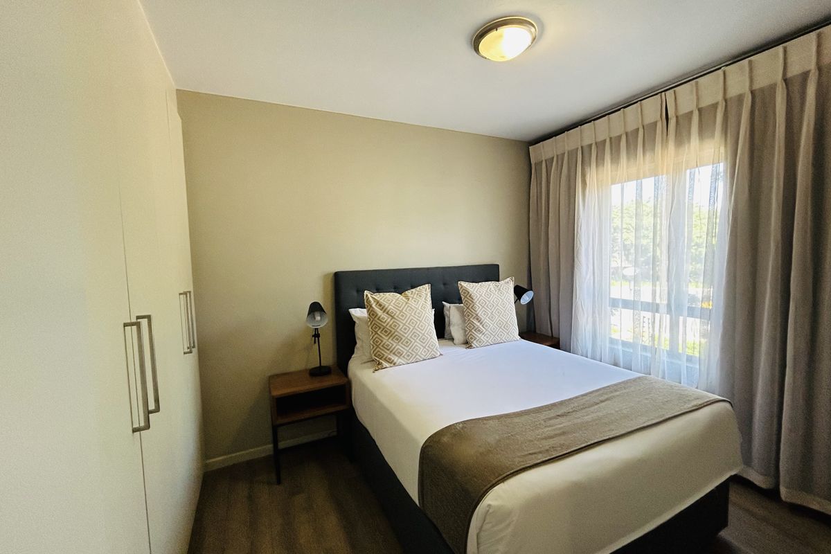 A well-maintained bedroom featuring built-in wardrobes, wood-look flooring, and large windows with curtains allowing for natural light.