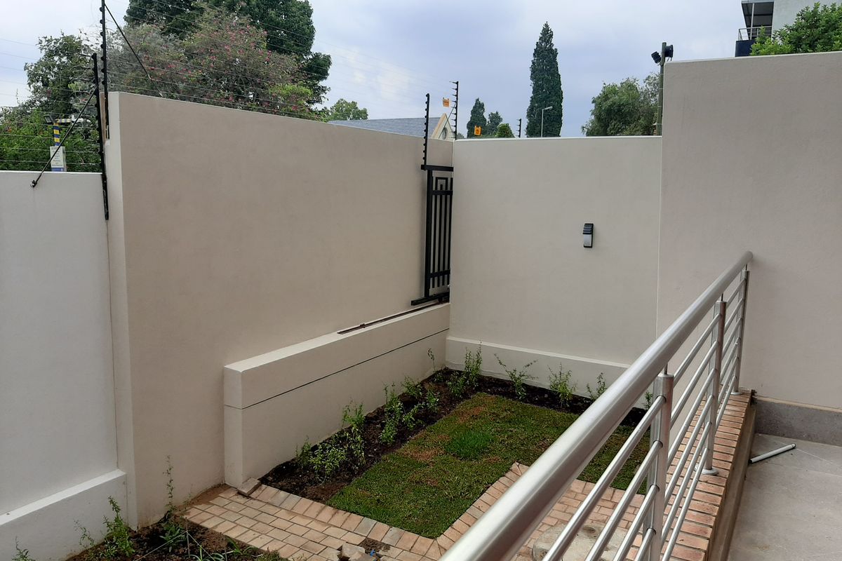 A private rear courtyard area featuring a paved walkway, small garden patch, secure boundary wall with electric fencing, and a modern stainless steel balustrade.