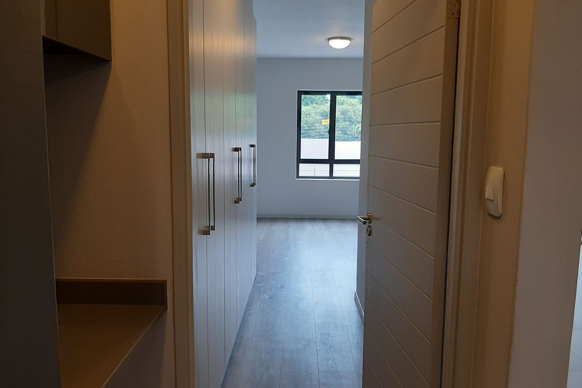 A bright, modern bedroom featuring built-in cupboards, laminate flooring, and a clean, neutral aesthetic with a view through the window.