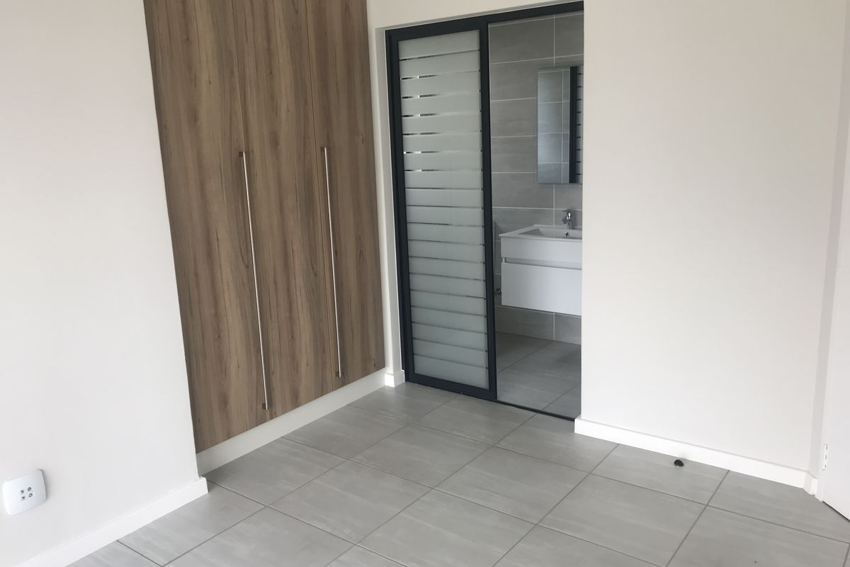 A modern, empty bedroom featuring neutral tiled flooring, built-in wood-finish cupboards, and a sliding door providing access to an en-suite bathroom.