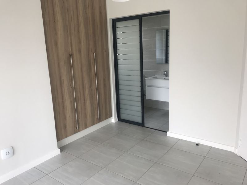 A modern, empty bedroom featuring neutral tiled flooring, built-in wood-finish cupboards, and a sliding door providing access to an en-suite bathroom.