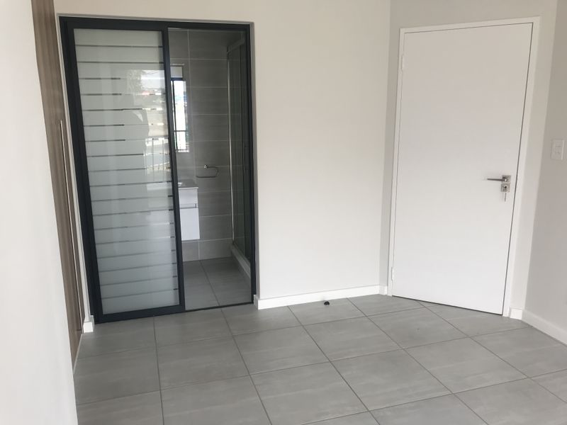 A clean, modern bedroom featuring neutral tiled flooring, a sleek sliding glass door leading to an en-suite, and a minimalist interior door.