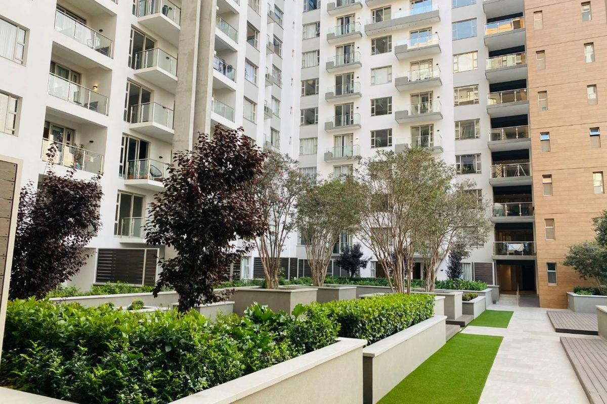 A well-maintained apartment complex courtyard featuring landscaped raised garden beds, artificial turf, paved pathways, and multiple levels of balconies with glass balustrades.