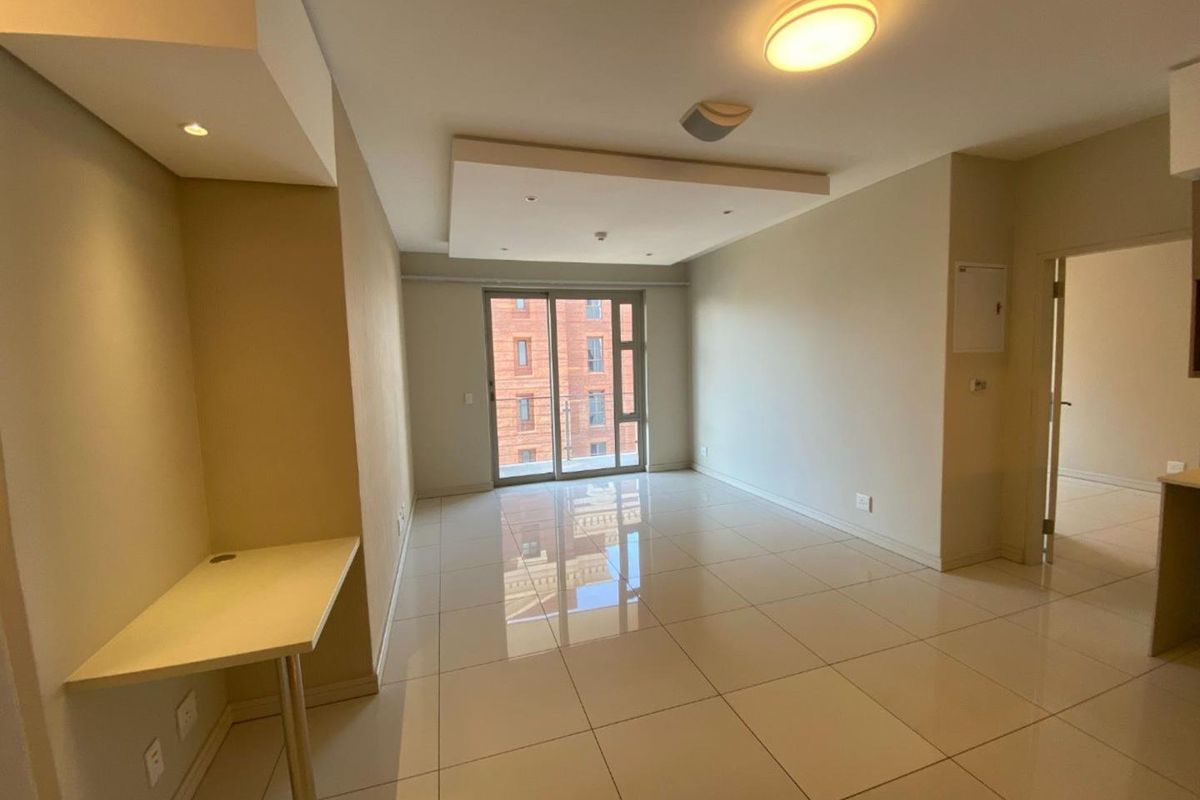 A bright, open-plan living area featuring polished tile flooring, a recessed ceiling with downlights, and a built-in desk, leading to a sliding glass door.