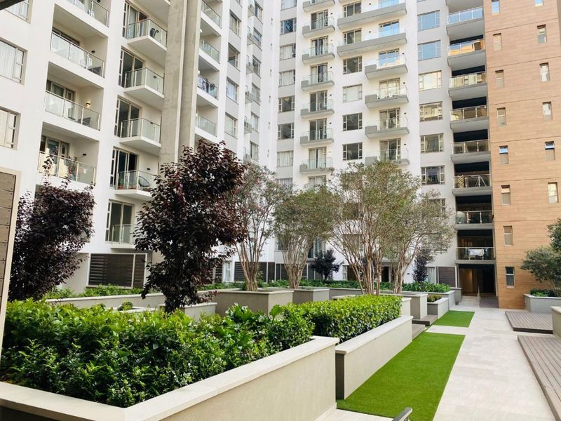 A well-maintained central courtyard area featuring landscaped planters, artificial turf, and paved walkways surrounded by a modern multi-story apartment building with private balconies.