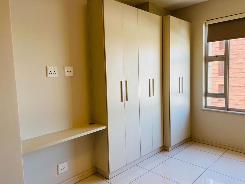 A bright bedroom featuring neutral tiled flooring, ample built-in cupboard storage, and a convenient integrated desk space with natural light.