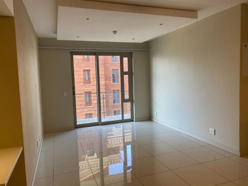 A bright, empty living room featuring polished tiled floors, a recessed ceiling with downlights, and a large sliding glass door leading to a balcony.
