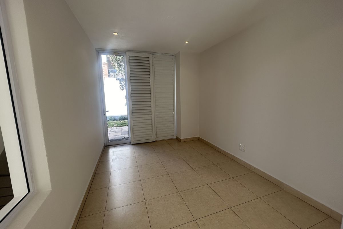 A bright, empty room featuring neutral floor tiles, recessed downlighting, and white plantation shutters opening to an outdoor area.