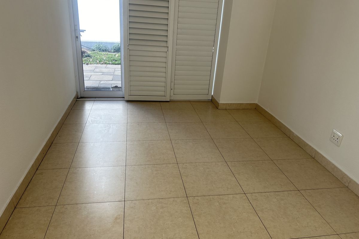 An empty room featuring neutral floor tiles and a glass door with white shutters, providing direct access to an outdoor patio area.