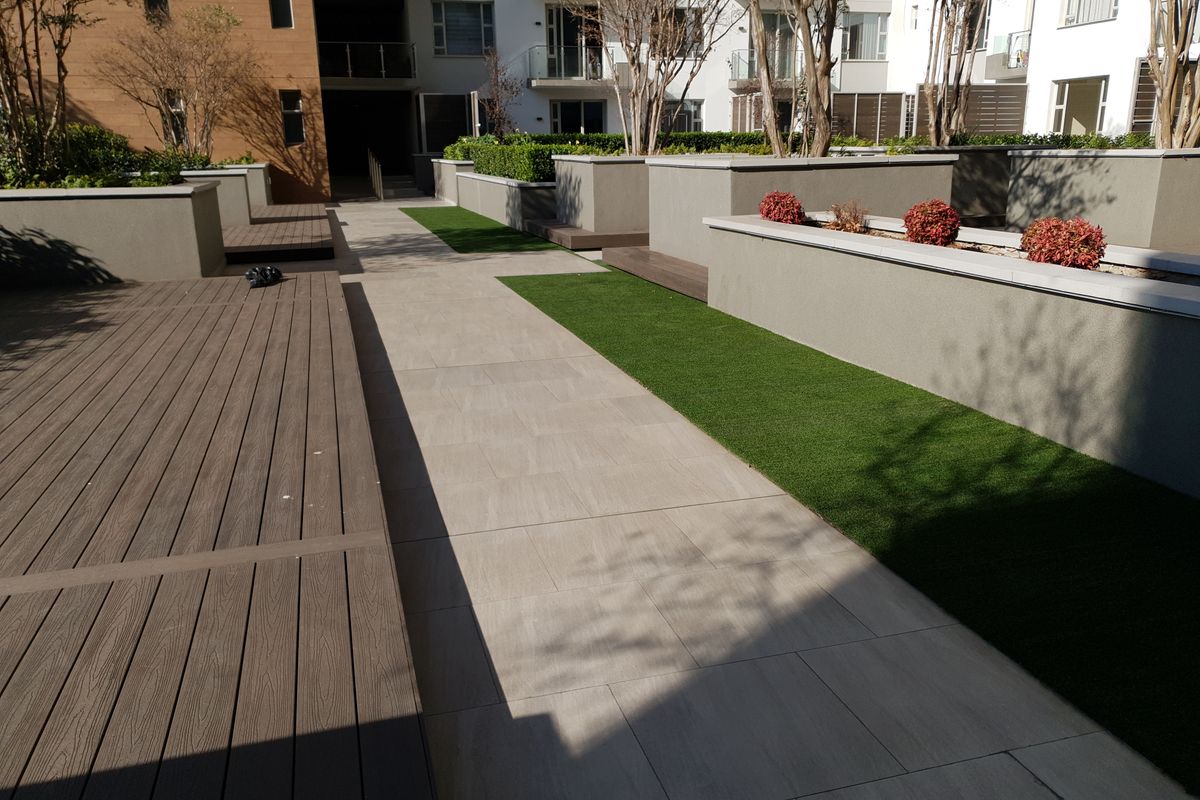 A well-maintained communal outdoor area featuring composite decking, tiled pathways, artificial turf, and modern raised planter boxes within an apartment complex.