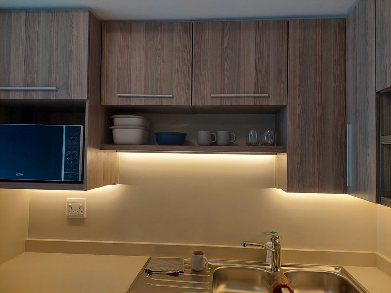 Modern kitchen area featuring wood-grain cabinetry, integrated microwave, stainless steel sink with mixer tap, and ambient under-cabinet LED lighting.