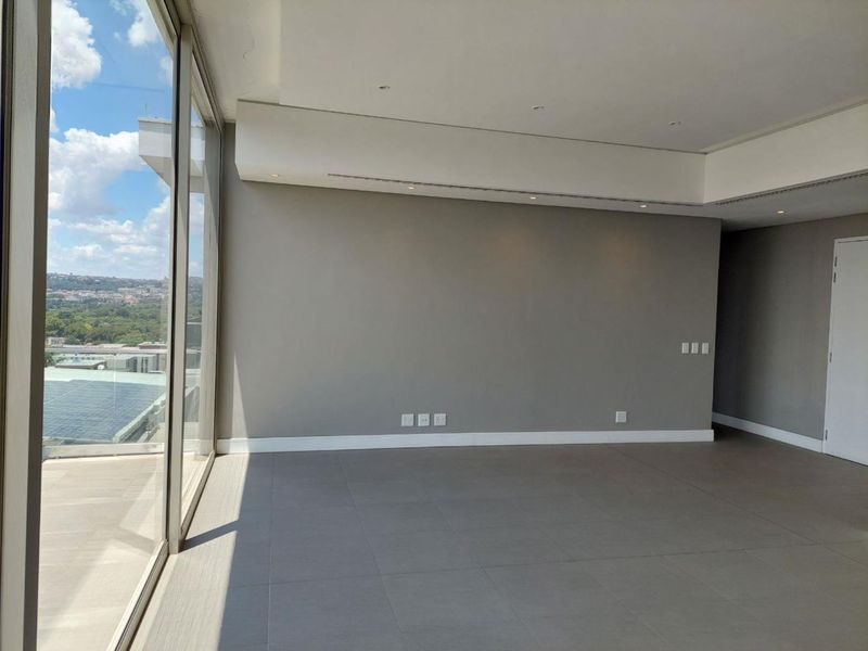 Spacious, modern living area featuring large floor-to-ceiling windows with expansive views, neutral grey walls, and clean tiled flooring.