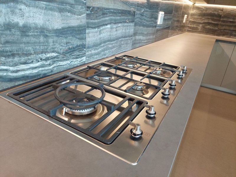 Modern kitchen featuring a high-end stainless steel gas cooktop set into a sleek stone countertop with a matching stone backsplash and integrated lighting.