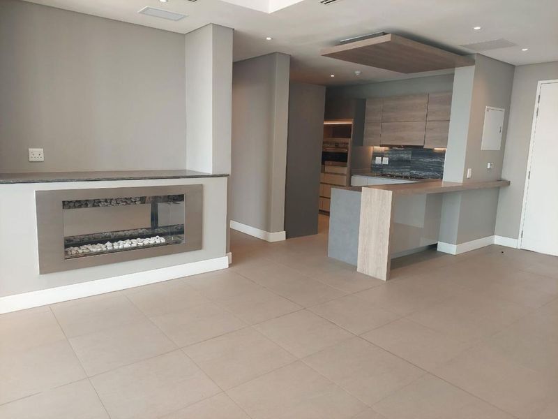 Modern open-plan living area featuring a sleek built-in fireplace, contemporary kitchen cabinetry, and a breakfast bar with neutral tiled flooring throughout.